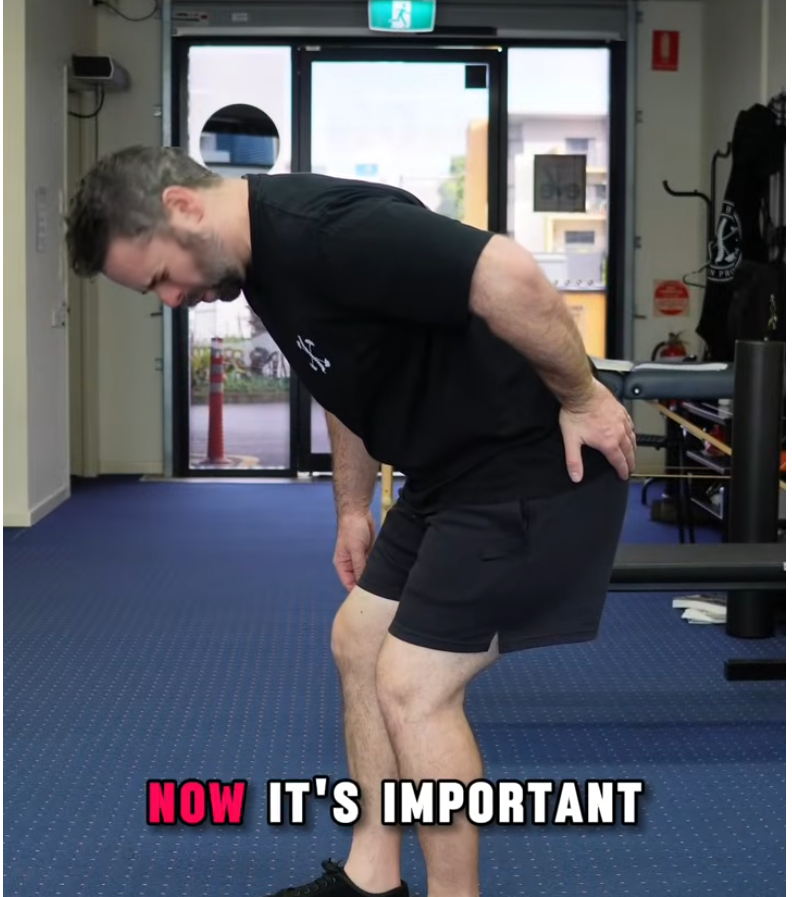 Man in a gym bends forward with hands on his lower back, wearing a black shirt and shorts; caption at bottom reads 'NOW IT'S IMPORTANT'