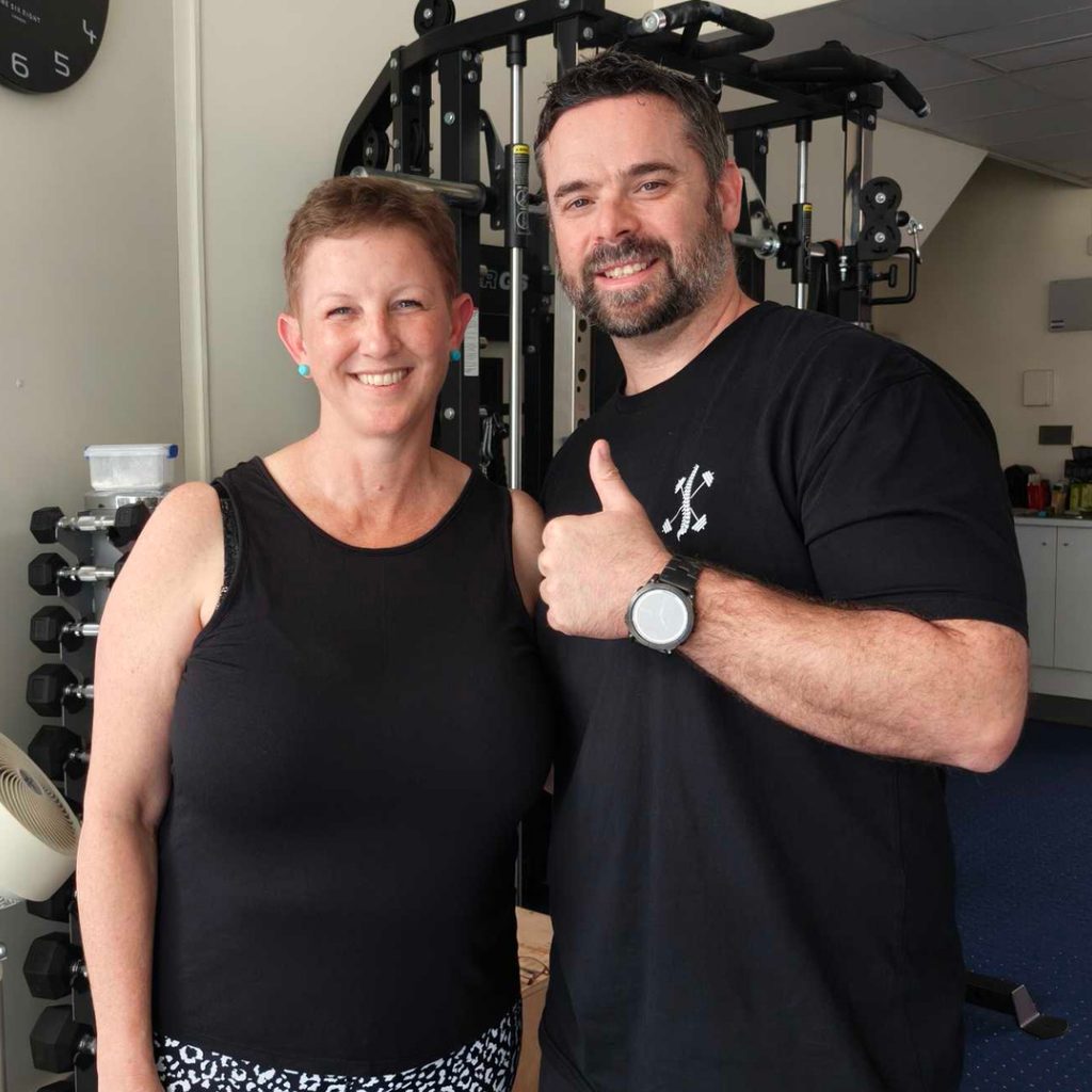 6a928bf9a41bab59d255292a-1787989059756-1 - Brisbane Back Pain Program | McGill Certified Practitioner | Online Coaching Two smiling adults in a gym posing by a weight machine, with one giving a thumbs-up.