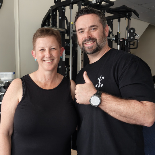 Susan 540 - Brisbane Back Pain Program | McGill Certified Practitioner | Online Coaching Two smiling adults in a gym posing for a photo with weight machines in the background.